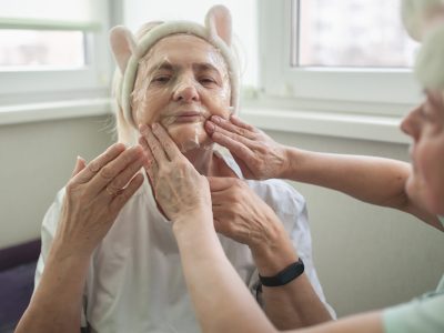 Two happy senior women apply facial cosmetic mask, have fun at home, body positivity, ageism A woman is getting her face waxed by two hands.